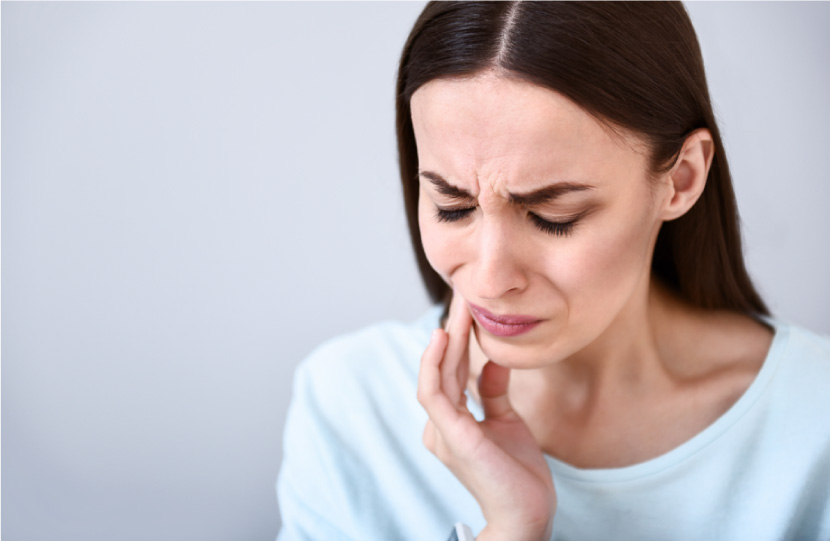 A brunette woman wearing a blue shirt cringes and touches her cheek in response to an intense toothache