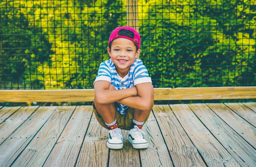 Smiling dark-haired boy wearing a red cap and striped blue shirt crouches on a wooden bridge in front of green trees