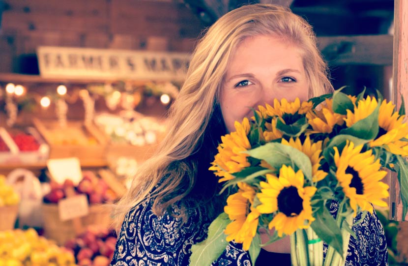 Blonde young woman hides her smile due to a chipped tooth with a bouquet of yellow sunflowers at a farmers market