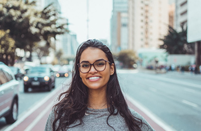 Brunette young woman wearing glasses smiles with veneers on a sidewalk in a busy city