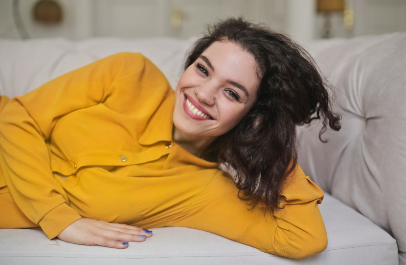 Brunette woman wearing a yellow blouse lies sideways on a tan couch smiling with no cavities or tooth decay
