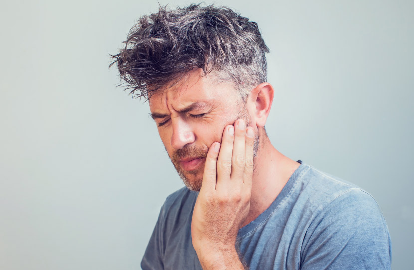 Middle-aged man in a gray shirt touches his jaw in pain because he grinds and clenches his teeth as he sleeps